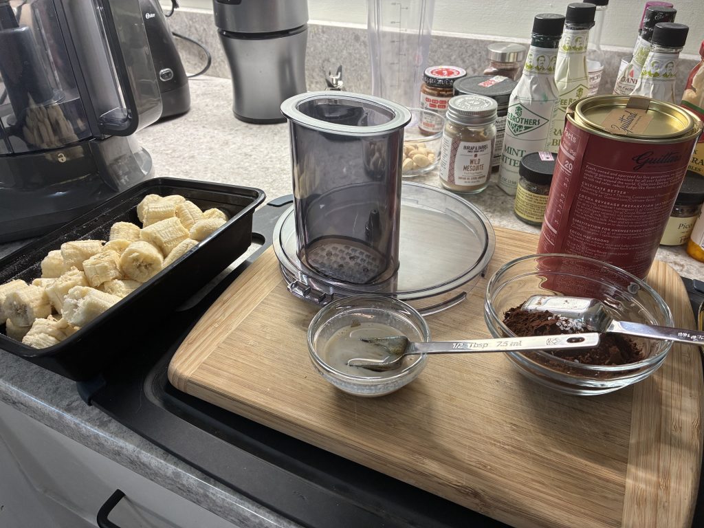 The fixing for nice cream on a kitchen counter. Chopped frozen bananas, half-and-half mixed with vanilla paste, cocoa powder.
